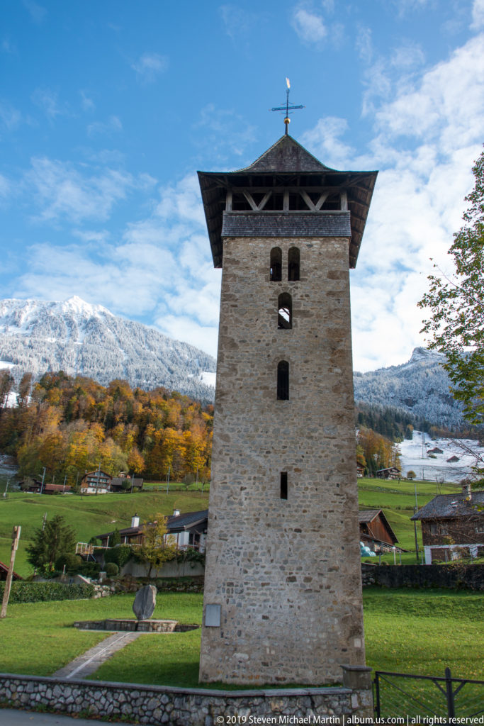 Square stone tower with a broad timber roof standing before snowy mountains and green fields.