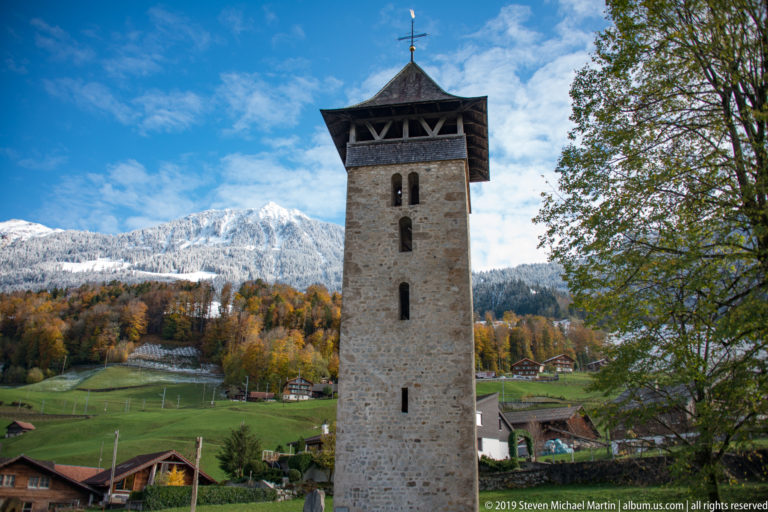 Tall stone tower with narrow openings and overhanging timber roof against a snowy mountain.