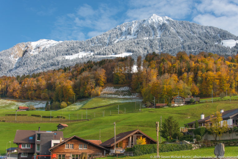 Alpine houses and pasture below autumn forest and a broad snow-covered mountain.