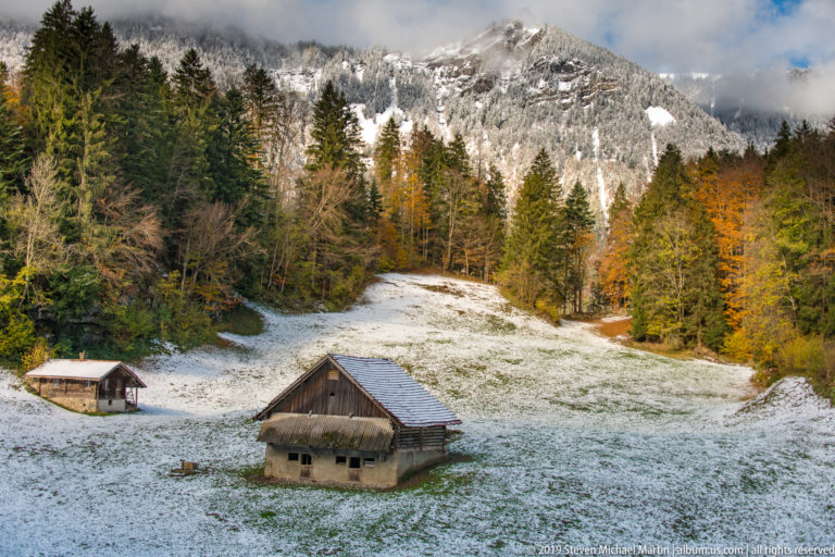 Two rustic barns in a lightly snow-covered meadow below forest and a rocky mountain.