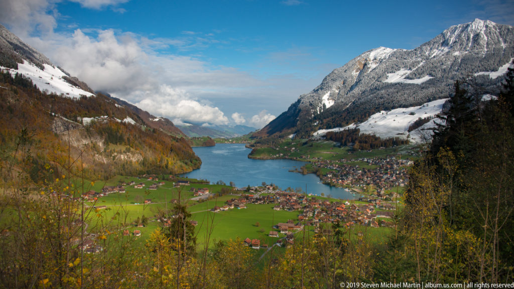 High view over a blue-green Alpine lake, lakeside village, fields, and snow-streaked mountains.