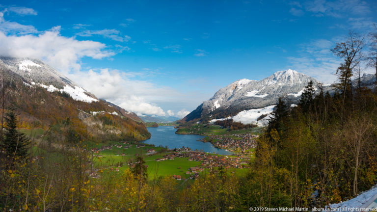 Wide view down an Alpine lake between mountains, with village roofs and green fields below.