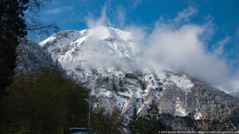 Snow-covered mountain face partly obscured by drifting white cloud above dark foreground trees.