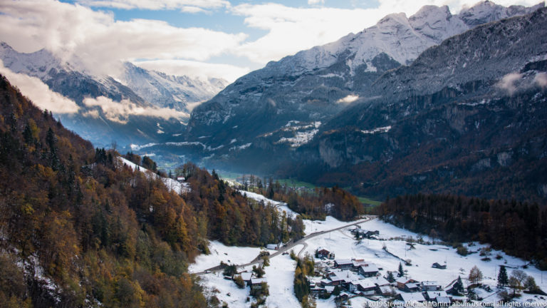 Village in a snowy Alpine valley surrounded by autumn forest, steep mountains, and low cloud.