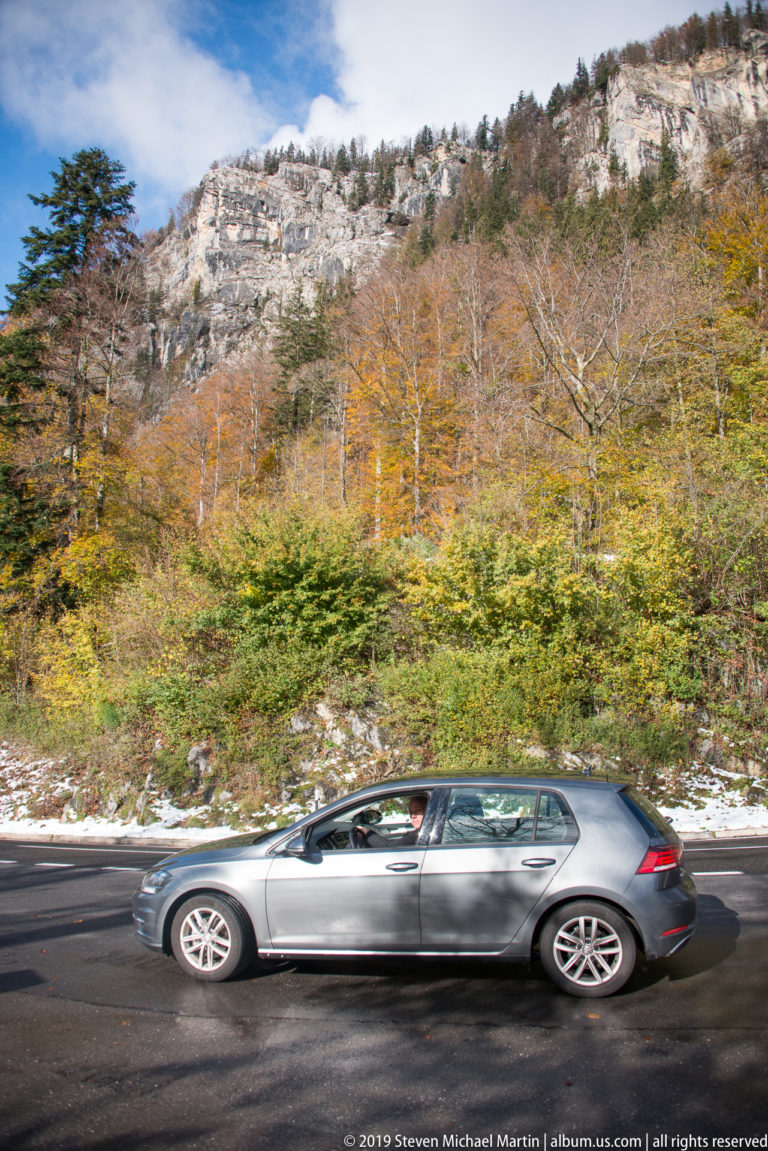 Silver station wagon driving along a mountain road beside snowy autumn woodland.