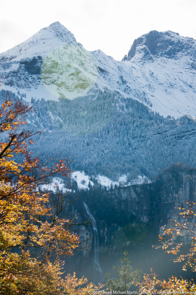 Dark mountain lake below snow-covered slopes, framed by orange autumn leaves.