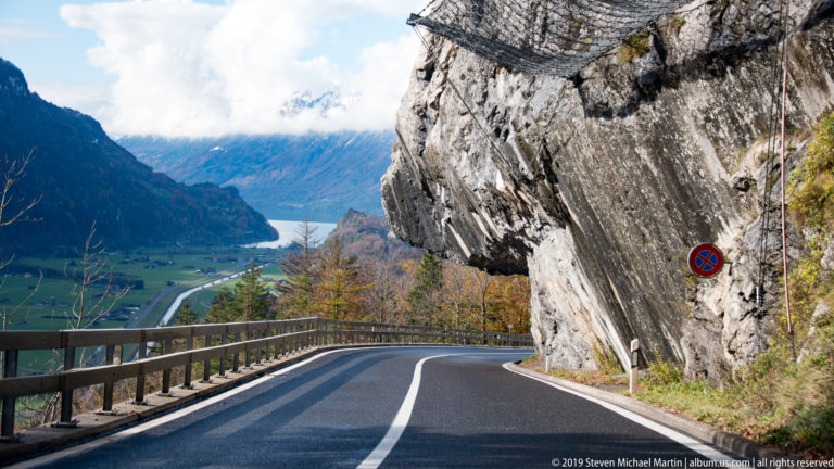 Two-lane road curves beneath a huge rock overhang above a blue Alpine lake and valley.