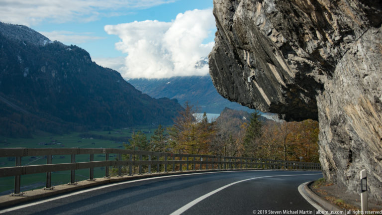 Cliffside road curves under projecting rock with a lake and valley visible ahead.
