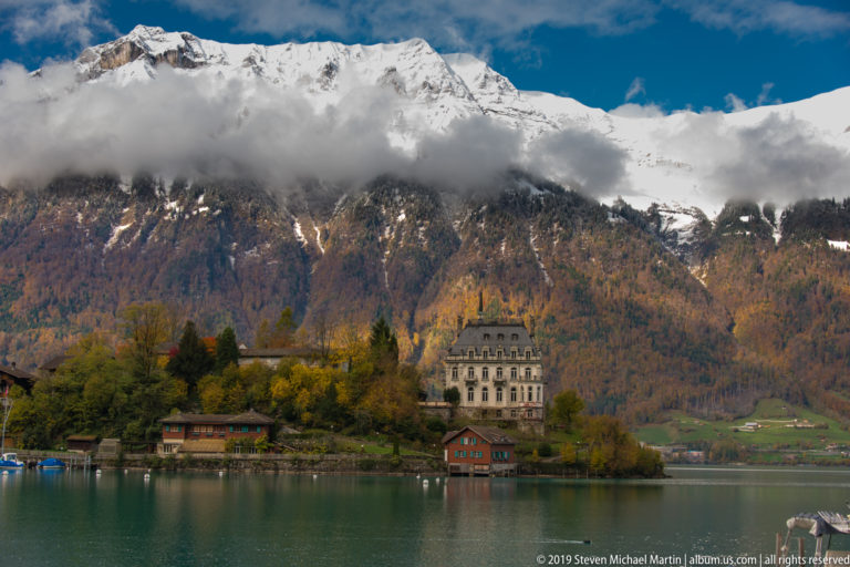 Large pale lakeside hotel with smaller timber buildings below snowy, cloud-wrapped mountains.