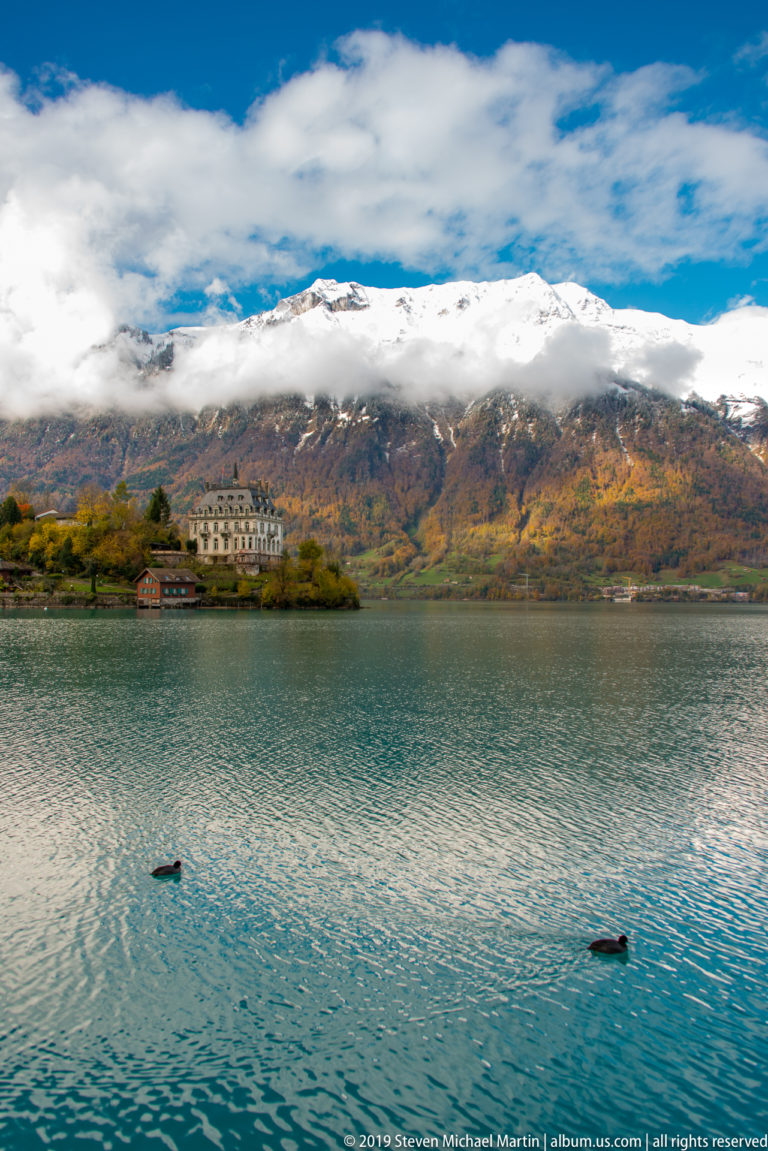 Pale hotel across a broad lake beneath snow-capped mountains, with two waterbirds in front.