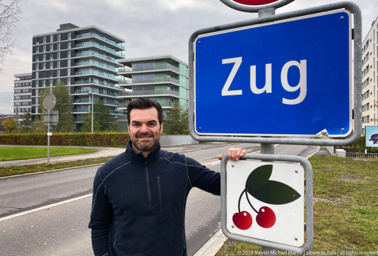 Smiling man holding a blue Zug sign above a cherry emblem, with modern apartment buildings behind.