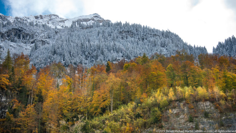 Orange and yellow autumn forest below a conifer ridge coated in fresh snow.