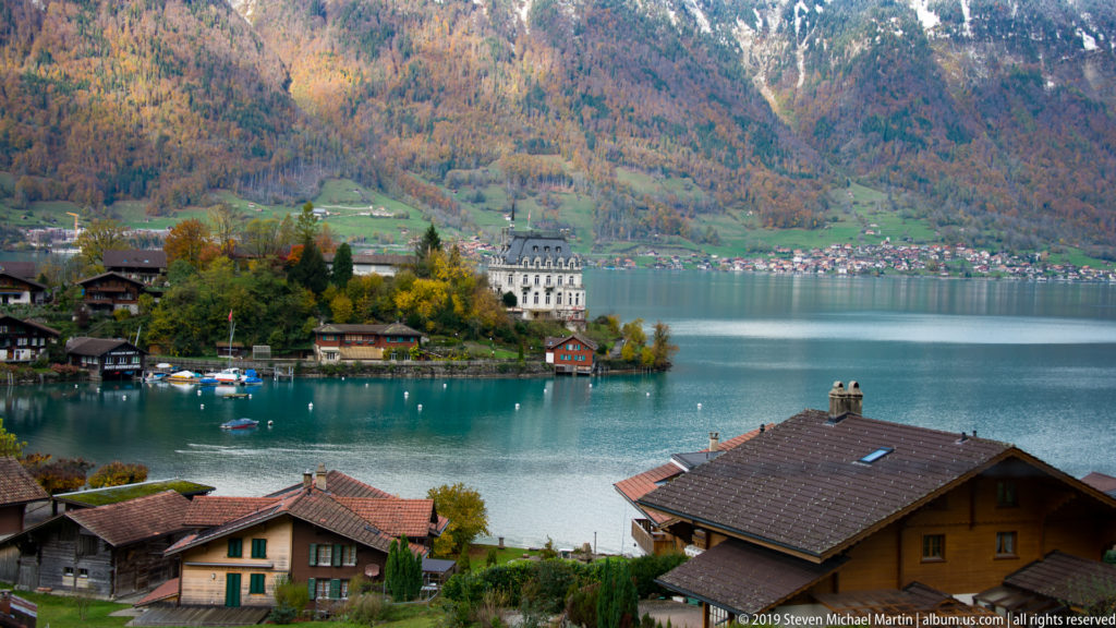Village rooftops descend toward a turquoise lake and a large pale hotel on a wooded point.