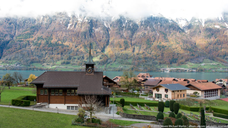 Small wooden church with clock tower and cemetery overlooking a lake and mountain village.