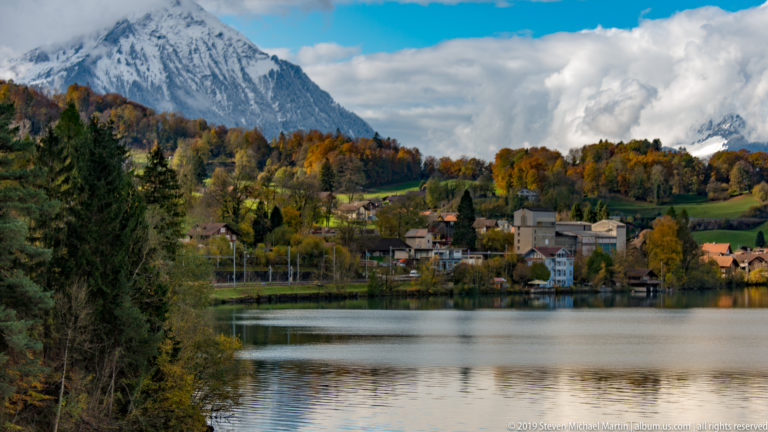 Calm Alpine lake facing a small shoreline town surrounded by golden autumn trees and mountains.
