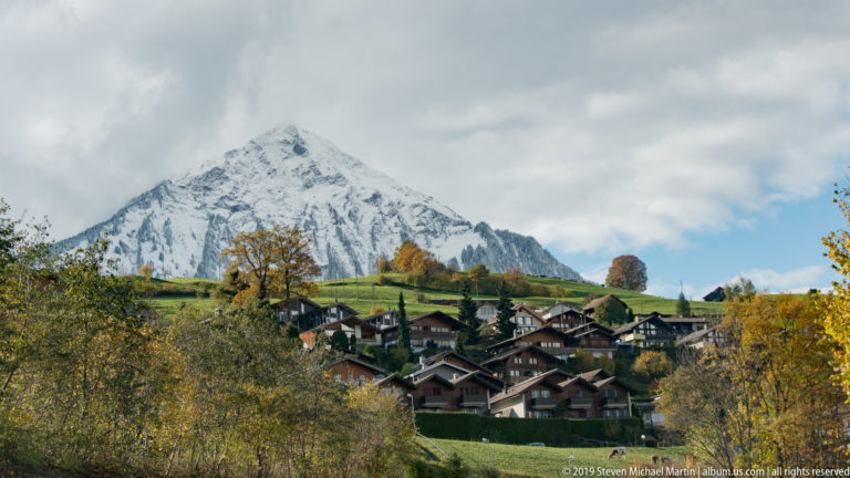 Chalet village on a green hillside beneath a steep pyramidal mountain covered with fresh snow.