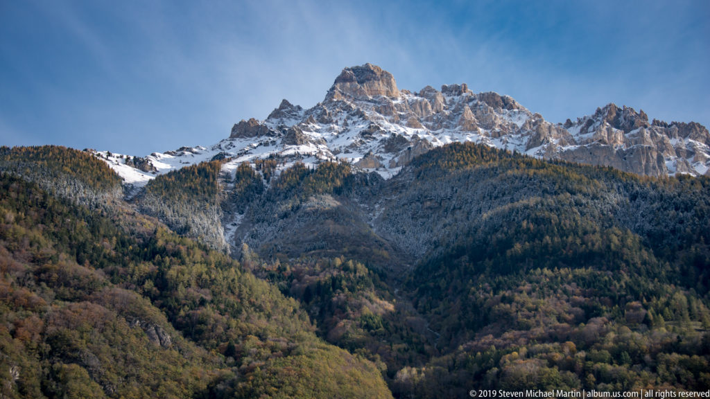 Jagged rocky Alpine summit dusted with snow above forested autumn slopes.