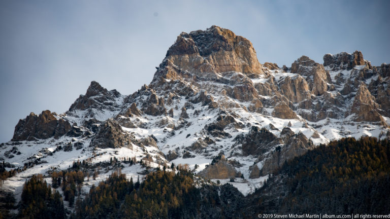 Jagged snow-dusted mountain cliffs above a dark conifer forest under a pale blue sky.