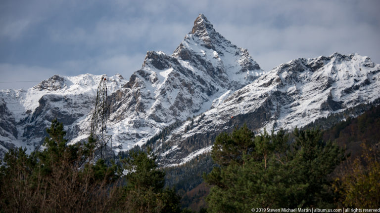 Snowy pointed mountain peak beyond forested slopes, with trees and utility lines in the foreground.