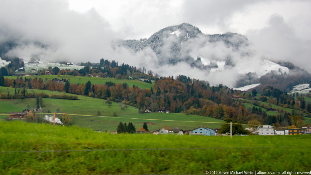 Green pastures, scattered houses, and autumn trees beneath low clouds covering wooded Swiss hills.