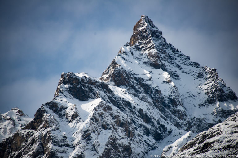 Steep pyramidal Alpine peak with snow in gullies and dark fractured rock faces beneath cloud.