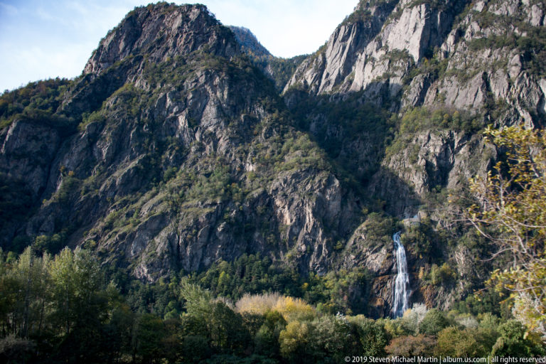 Slender waterfall dropping down a steep rocky cliff above mixed autumn woodland.