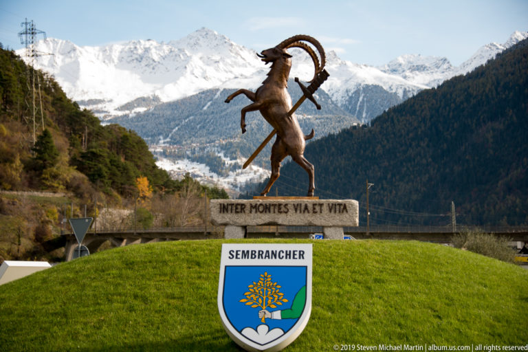 Ibex sculpture above a Sembrancher shield and Latin inscription, with snowy mountains across the valley.