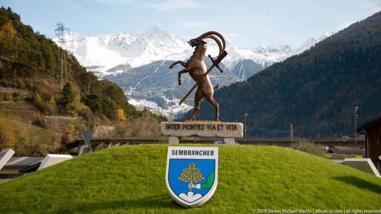 Rearing ibex monument above the Sembrancher coat of arms, backed by a forested valley and snowy peaks.