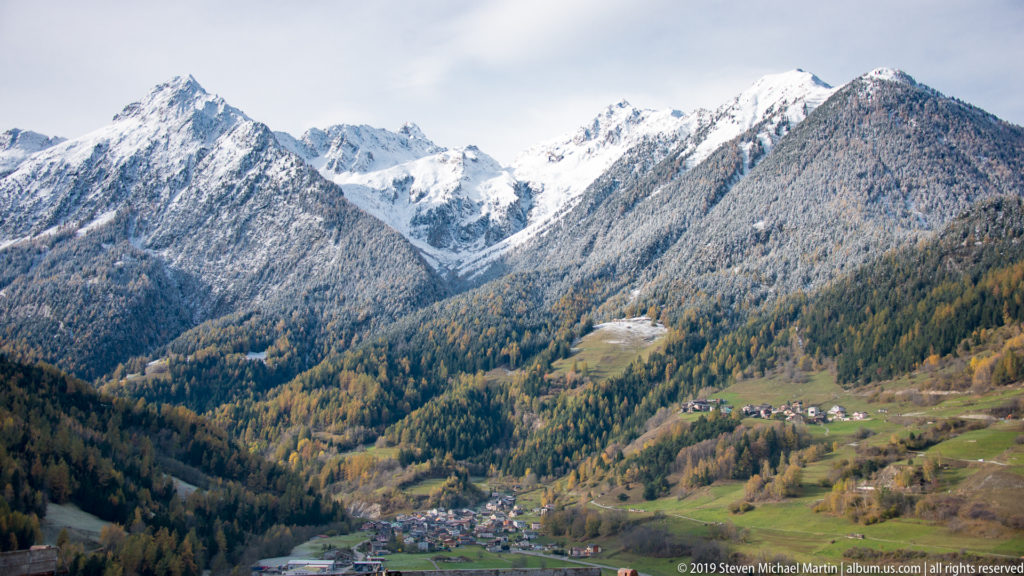Village and green-brown pastures in an Alpine valley beneath snow-covered forested mountains.