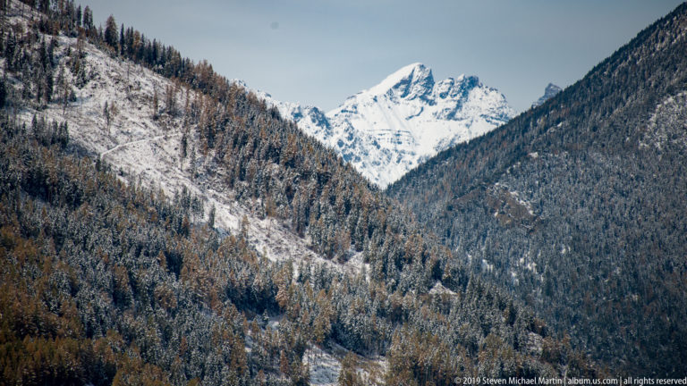 Snow-covered mountain peak centered beyond a V-shaped valley of forested, lightly snow-dusted slopes.