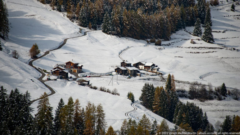 Small cluster of Alpine houses and farm buildings surrounded by snowy fields, trees, and a winding road.
