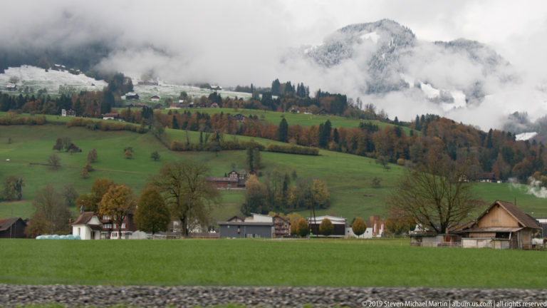 Green field and rural houses below patchwork autumn hills partly hidden by low cloud.