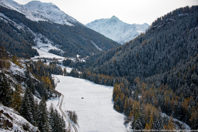 Broad snow-covered valley floor with a track, forested slopes, and a distant white mountain.