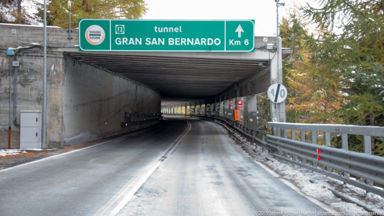 Road entering a covered approach beneath a green “tunnel GRAN SAN BERNARDO Km 6” sign.