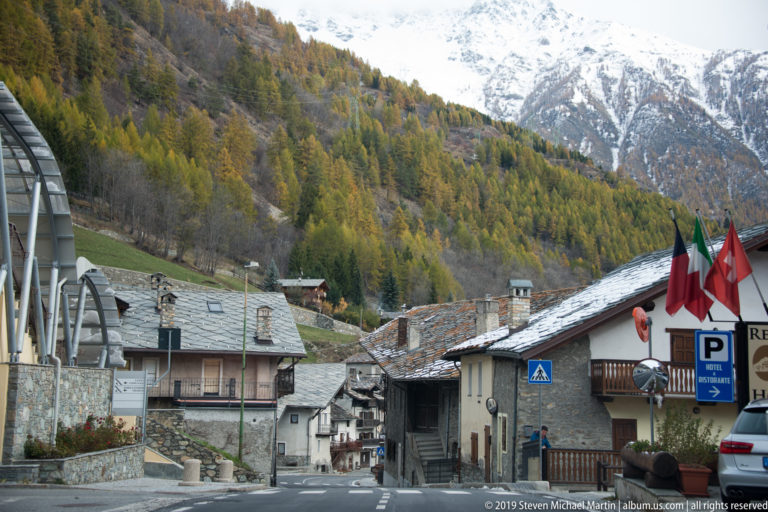 Narrow Alpine village street with stone-roofed houses, Italian and Swiss flags, larch forest, and snowy mountains.