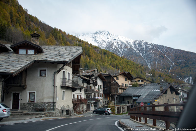 Curving Alpine village street lined with traditional houses, with autumn forest and a snow-covered mountain behind.