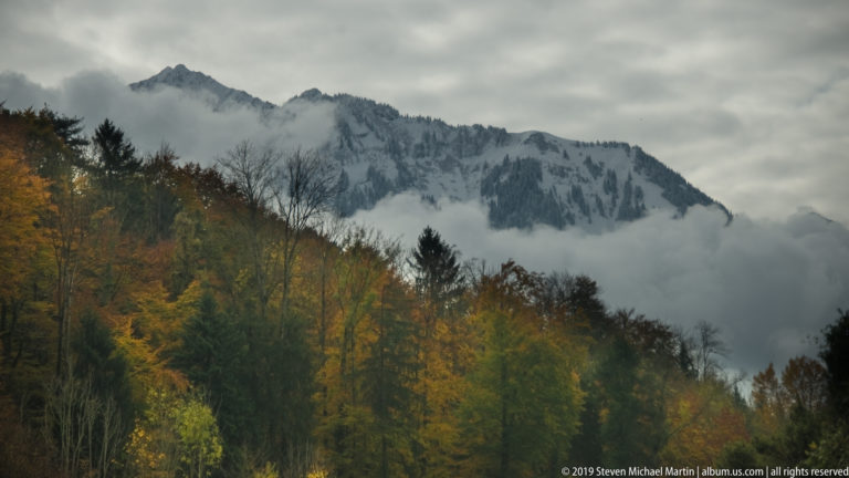 Golden autumn forest below a pale rocky mountain ridge partly obscured by cloud.