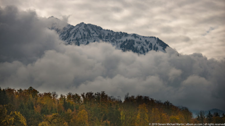Jagged gray mountain ridge emerging from heavy cloud above a band of autumn-colored treetops.
