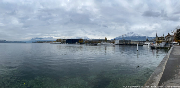 Lake Lucerne waterfront under clouds, with the KKL cultural complex, city shoreline, and snow-dusted mountains in the distance.