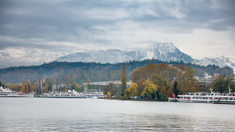 Lake Lucerne with passenger boats, autumn trees, wooded ridge, and a pale mountain beneath an overcast sky.