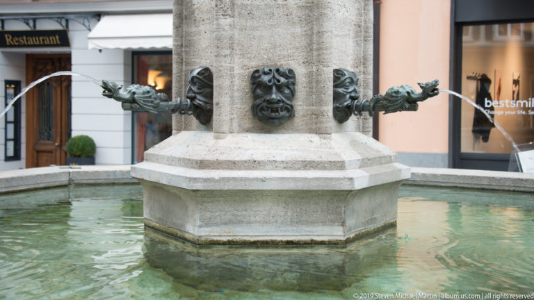Stone fountain column with carved face and several bronze spouts pouring water into a basin.