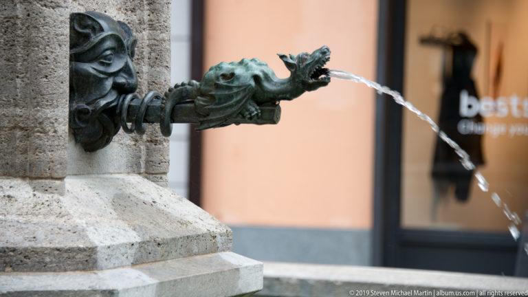 Close view of a bronze dragon-headed fountain spout pouring water from its mouth.