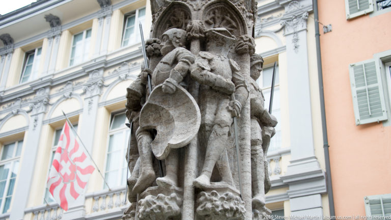 Carved stone figure in historic dress holding a shield atop a Lucerne fountain column.