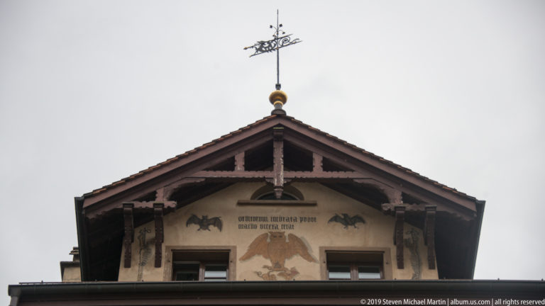 Historic gable with painted birds and inscription beneath timber framing, topped by an ornate weather vane.