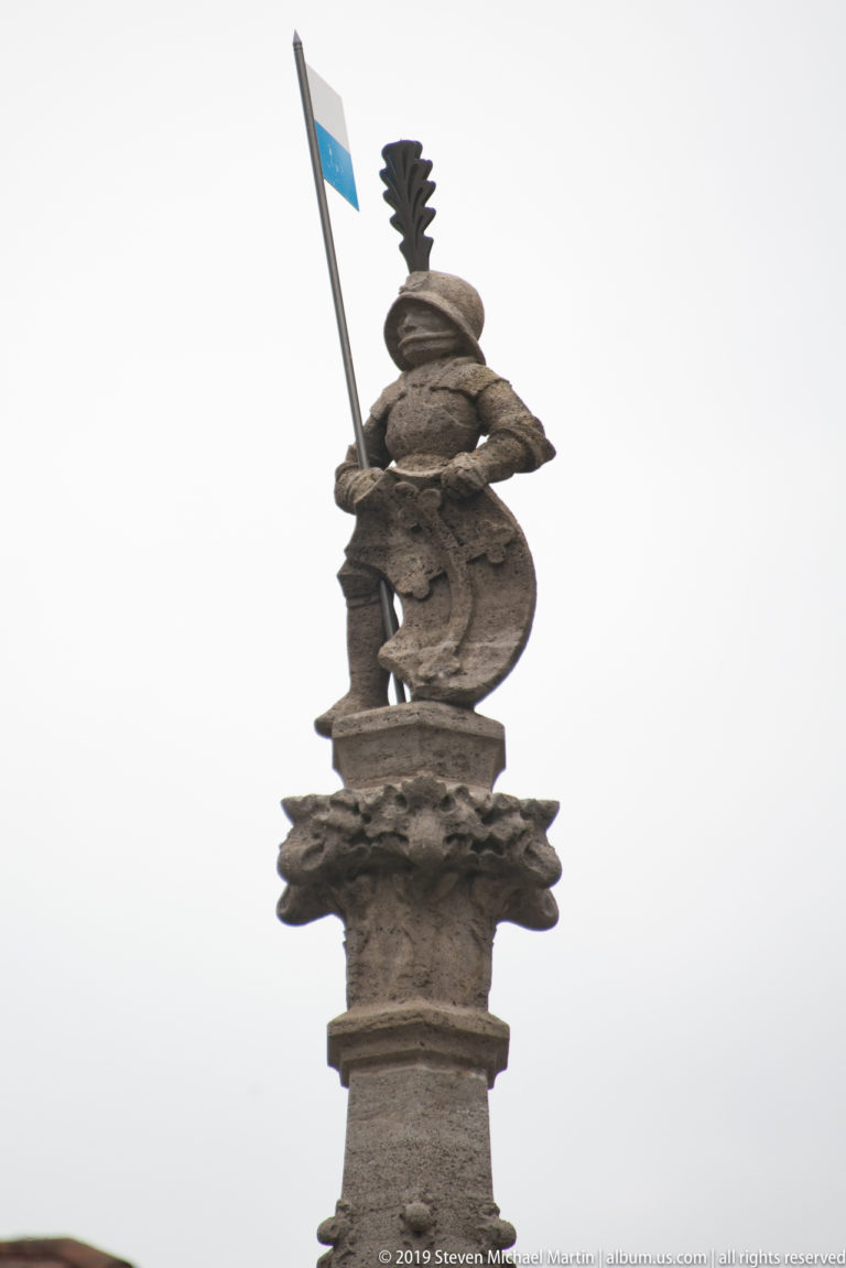 Carved figure atop a fountain holding a blue-and-white banner against the sky.