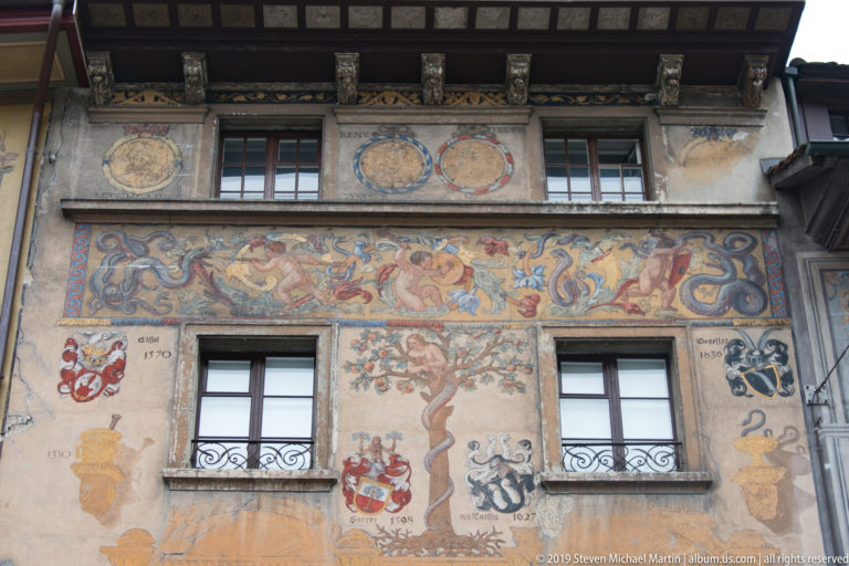 Painted Lucerne façade detail with heraldic shields, central tree motif, windows, and colorful ornamental frieze.