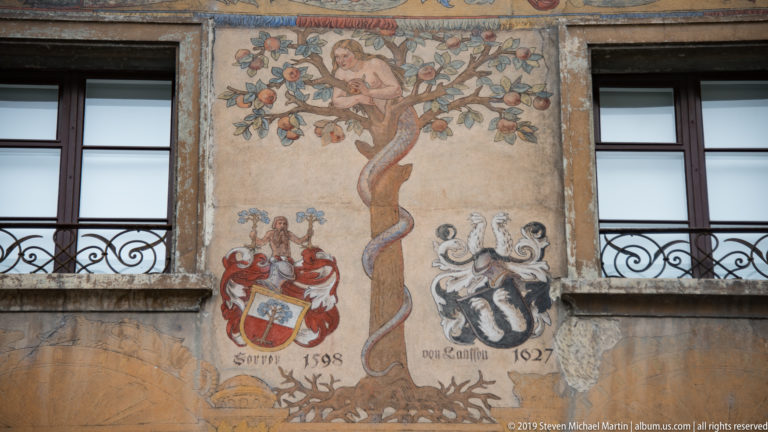 Painted tree and two heraldic shields with dates between windows on a historic Lucerne façade.