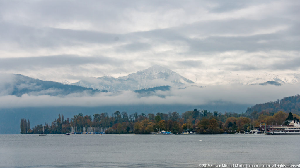 Mist and low cloud layered across mountains beyond Lake Lucerne, with a dark tree-lined shore in the foreground.