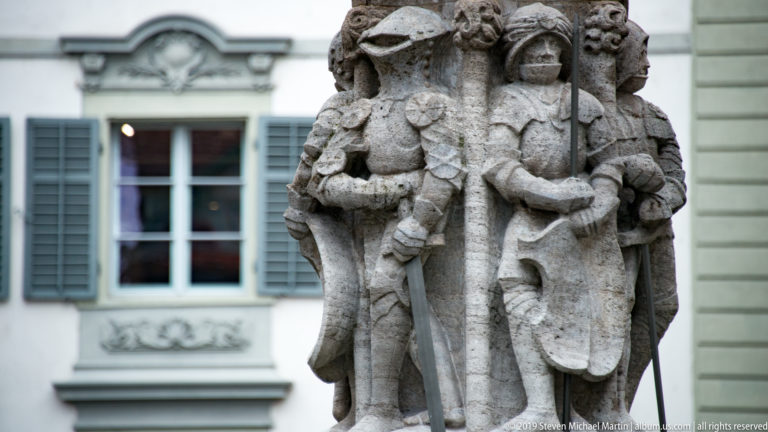 Group of weathered carved stone figures in historic dress standing together on a Lucerne fountain monument.