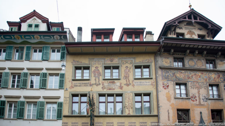 Adjacent historic Lucerne façades with figure murals, heraldic decoration, inscriptions, and neighboring green shutters.